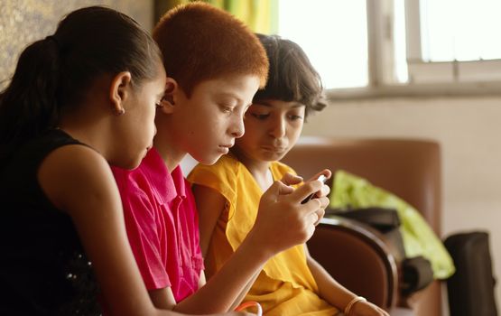 Three multi ethnic kids or siblings busy in playing games on mobile at home - concept of childrens mobile video game addiction, using technology, internet on smartphone