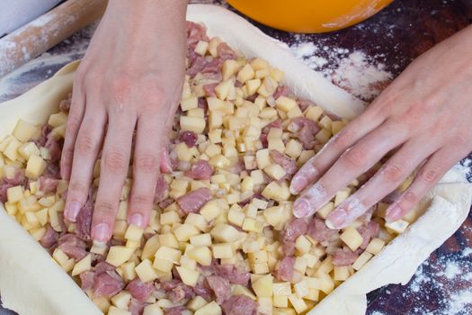 Cooking pie with potatoes and meat. Women's hands lay out cut potatoes and pieces of meat on the rolled dough lying on the table.