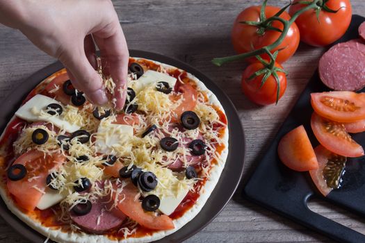 Hands sprinkle cheese on pizza. Pizza with tomatoes, olives and cheese is on the plate. Ingredients for cooking pizza are on a wooden table. Cooking pizza at home.