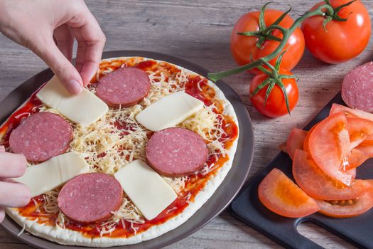 Hands sprinkle the pizza with cheese. On a plate-pizza with tomatoes, bacon, sausage and cheese. The ingredients for making pizza are on a wooden table. Cooking pizza at home.