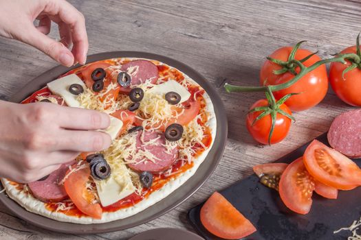 Hands sprinkle the pizza with cheese. On a plate-pizza with tomatoes, olives, bacon, sausage and cheese. The ingredients for making pizza are on a wooden table. Cooking pizza at home.