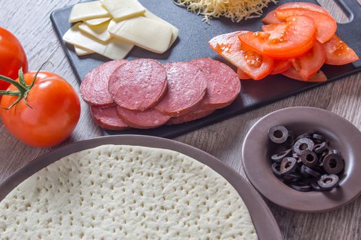 Pizza base and ingredients for its preparation. Tomatoes, sausage, cheese and olives are on the kitchen table. Cooking pizza at home.