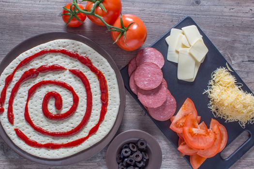 Cooking pizza at home. Pizza base and ingredients for its preparation. Tomatoes, sausage, cheese and olives are on the kitchen table.