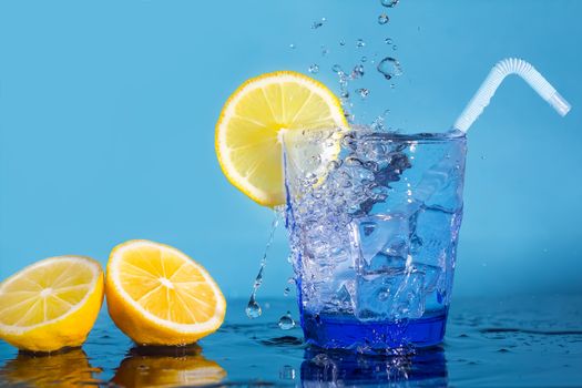 A glass with water and ice cubes,a slice of fresh lemon,a tube and a spray of water falling from above. Drops of water fall on a fresh lemon. A glass with a drink stands on a table with sliced lemons