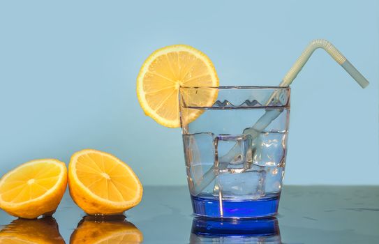 A glass of water, ice, lemon, and straw sits on a table with a blue background. Preparing a fresh drink with lemon juice. Sliced yellow lemons lie on the table next to a glass of water.Making soda