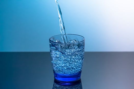 Clean water is poured into a glass standing on a black table. Blue glass with flowing water standing on a black table on a blue background