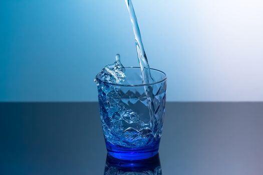 Clean water is poured into a glass standing on a black table. Blue glass with flowing water standing on a black table on a blue background