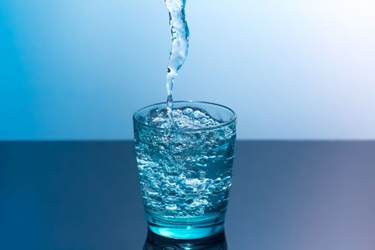 Blue glass with flowing water standing on a black table on a blue background. Clean water is poured into a glass standing on a black table.