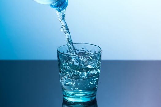 In a glass standing on a black table pour clean water from a bottle. Blue glass with flowing water standing on a black table on a blue background