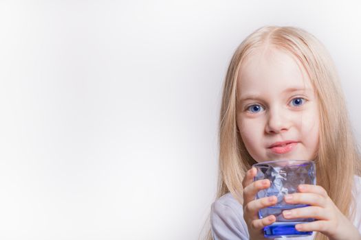 A child girl of Caucasian appearance holds a glass of water in her hand close-up on a white background with a place for the inscription. A blonde girl with long hair and blue eyes drinks water