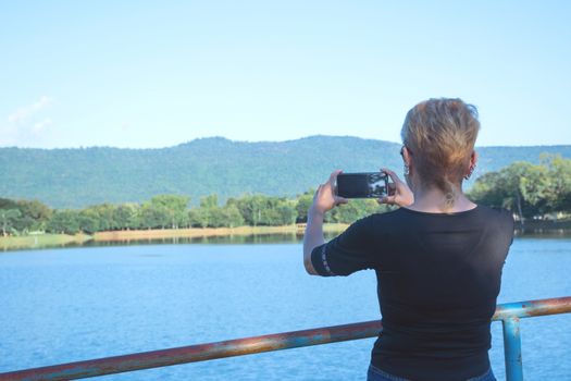 Woman making photo with smart phone. Happy traveling female making photo on mountains and river background.