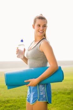 Sporty blonde holding exercise mat and water bottle in parkland