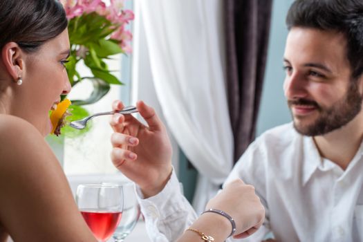 Close up portrait of young man feeding girlfriend with fork at dinner.