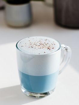 Blue pea latte or blue matcha latte with copy space. Hot fresh milk with blue butterfly pea flowers or clitoria ternatea latte in glass cup at natural sunset hard light. White background. Vertical