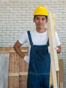 An Asian carpenter wearing a yellow hardhat, carrying wood for making furniture in the workshop room.