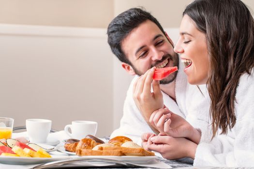 Close up fun portrait of couple in bathrobe on bed enjoying breakfast together. 