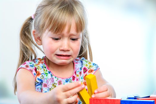 Close up portrait of crying little girl playing with wooden blocks at table.Frustrated girl showing moody behavior and long face.
