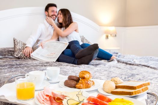 Close up detail of breakfast tray on bed with laughing young couple in background.