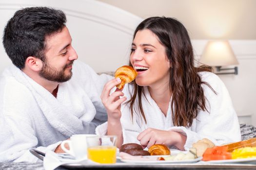 Close up portrait of couple having fun at breakfast in hotel room.Girl about to take bite of croissant.
