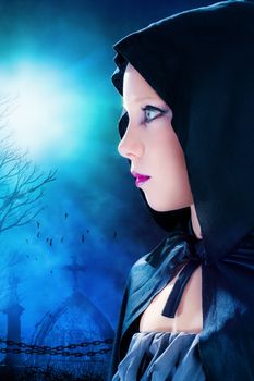 Close up portrait of attractive young mysterious gothic girl wearing black hood.Graveyard with full moon in background.