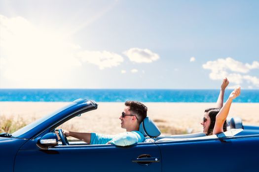 Close up portrait of young man driving blue convertible. Girlfriend sitting in back raising arms in air.