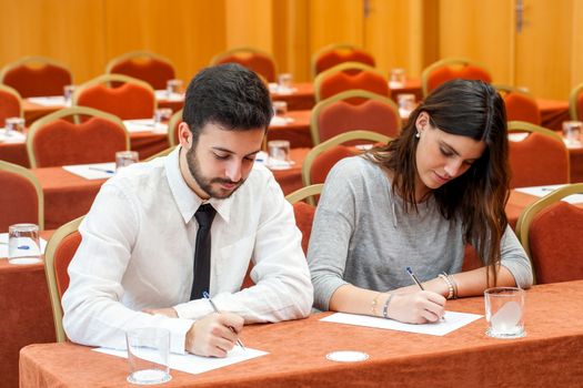 Portrait of young business couple taking notes in conference room.Couple sitting at table writing with pens with rows of seats in background.