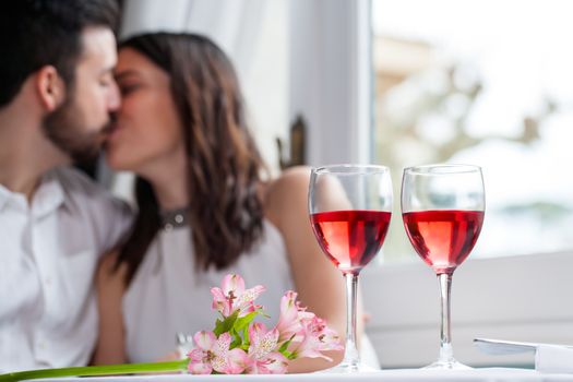 Close up detail of two wineglasses with couple in background kissing. Wine glasses filled with rose wine next to flower bouquet.