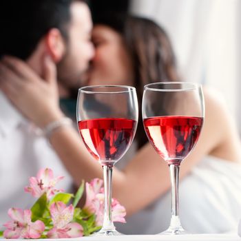 Close up of two wineglasses with rose wine and flower bouquet.In background a out of focus couple kissing.