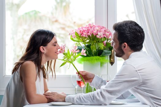 Close up portrait of young man giving flower to girlfriend in restaurant. Elegant couple in white casual wear sitting next to window.