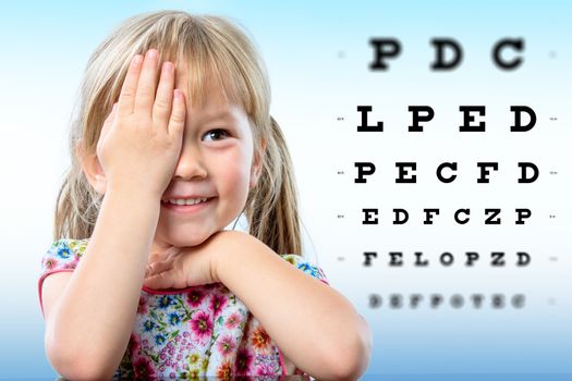 Cute little girl reviewing eyesight.Girl closing one eye with hand reading block letters on vision chart with focus point.