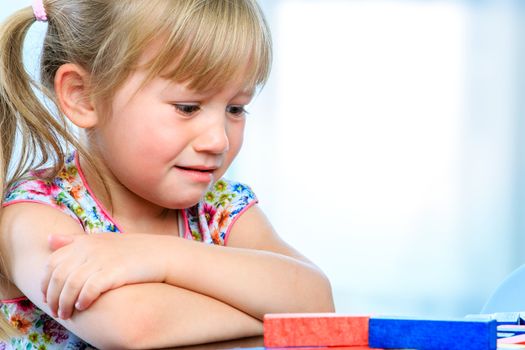 Close up portrait of unhappy little girl at table with educational game. Infant showing miserable sad face expression.