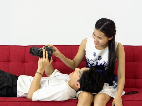 Young lovers spend time together on holidays in the photo studio. The young man allow her girlfriend to view pictures taken on the camera screen while relaxing on a red sofa.