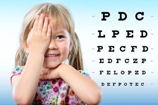 Close up portrait of cute little infant reviewing eyesight on chart. Girl closing one eye with hand reading vision chart.