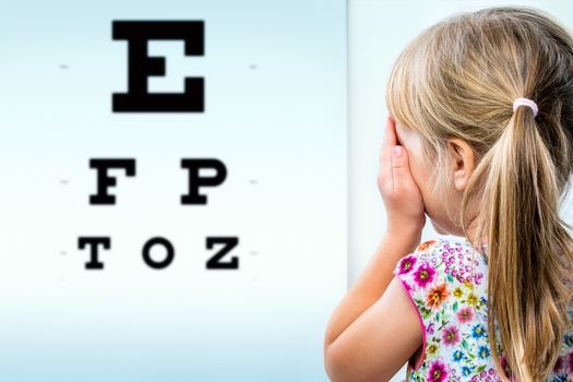 Close up rear view of girl testing eyesight. Infant closing one eye with hand looking at vision test chart.