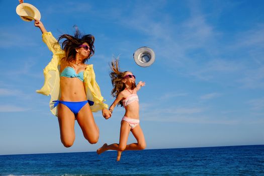 Action portrait of young mother with daughter  jumping together on beach. Laughing girls in swimwear throwing hats in air.