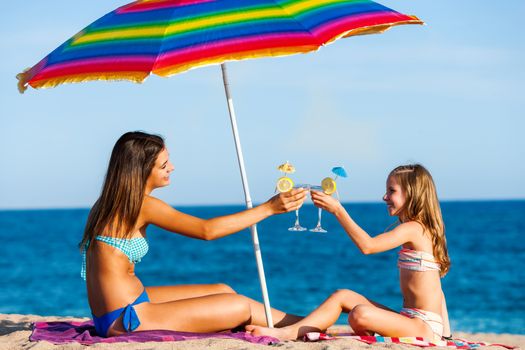 Portrait of two young girls on summer holiday. Young women sitting under colorful umbrella on beach drinking fruit cocktails.