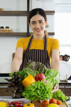 Young housewife smiled and offered a basket full of various kinds of vegetables on the front. Morning atmosphere in a modern kitchen.