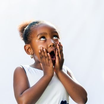 Portrait of surprised little african girl with hands on face looking up.Isolated against light background.