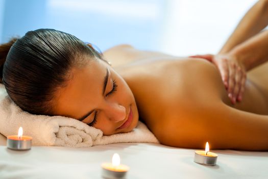 Close up portrait of attractive young woman having relaxing body spa treatment in dim candle light. Therapist in background massaging back.