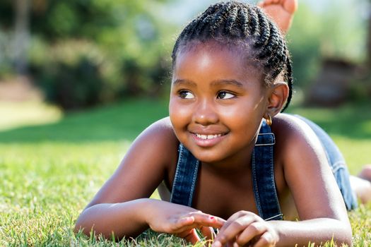Close up portrait of cute little african girl with braids laying on green grass.
