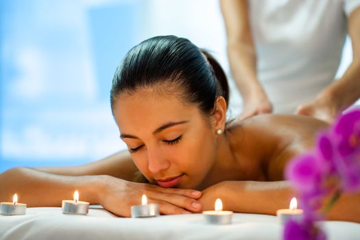 Close up portrait of young woman relaxing in dime ambient light with candles in spa. Therapist in background massaging back.