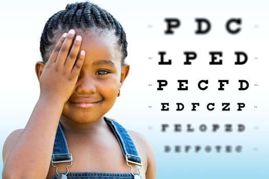 Close up Face shot of little African girl testing vision. Girl with braided hairstyle closing on eye with hand. Vision chart with block letters and focus point in background.
