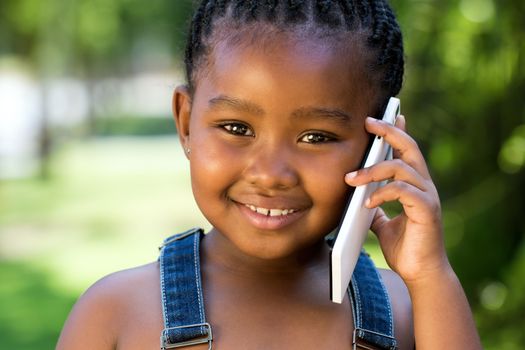 Close up face shot of cute little african girl talking on smart phone against green outdoor background.