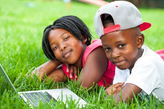 Close up portrait of African boy and girl laying on green grass with laptop.