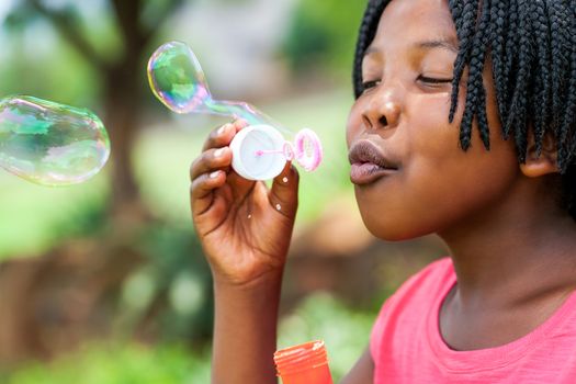 Close up portrait of cute African girl with braids blowing bubbles in park.