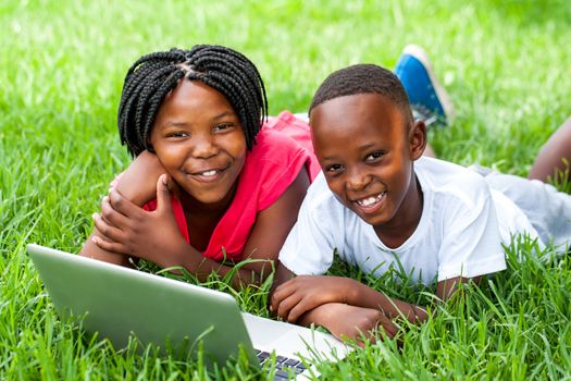 Close up portrait of cute African kids laying on grass with laptop.