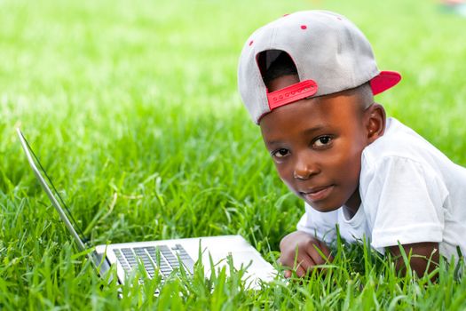 Close up portrait of African boy with baseball hat laying with laptop on grass.
