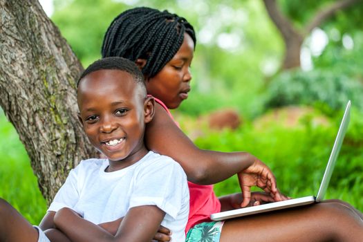 Close up portrait of happy African kids under tree playing on laptop.