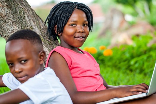 Close up portrait of cute african girl typing on laptop next to brother under tree in park.