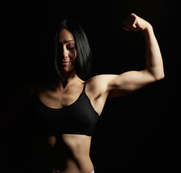 young beautiful athletic girl raised and bent her arm demonstrating her biceps, athlete is standing on a dark background, wearing a black top and short shorts